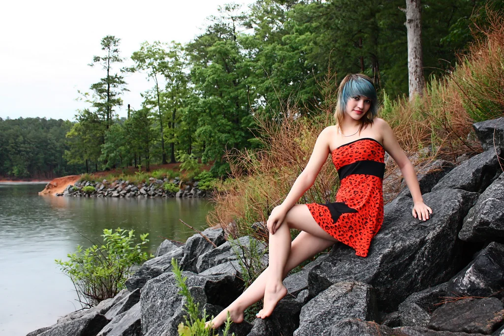 A beautiful young woman posing in a red dress on rocks by a lake : Free Stock Photo